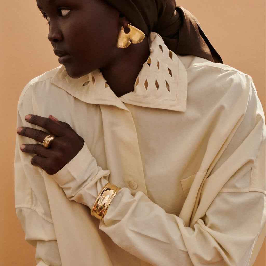 Woman wearing a beige coat with decorative collar, gold jewelry, and a brown headscarf against a beige background