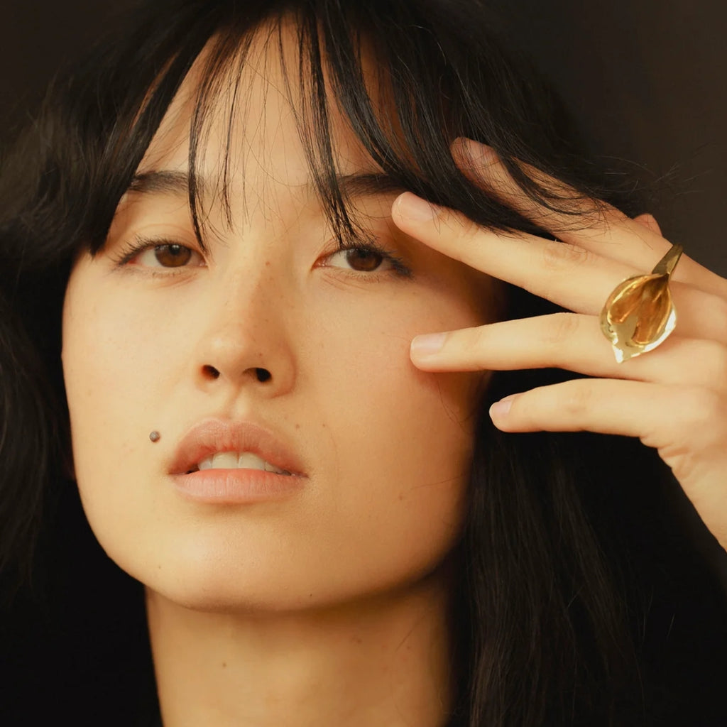 Woman with dark hair and a gold ring on her finger against a dark background