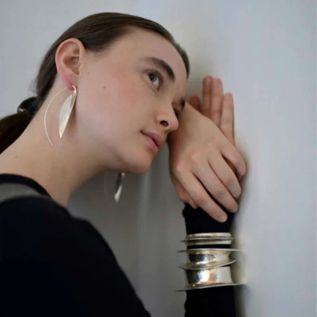 Woman wearing large hoop earrings and a black top with silver jewelry, leaning against a white wall.