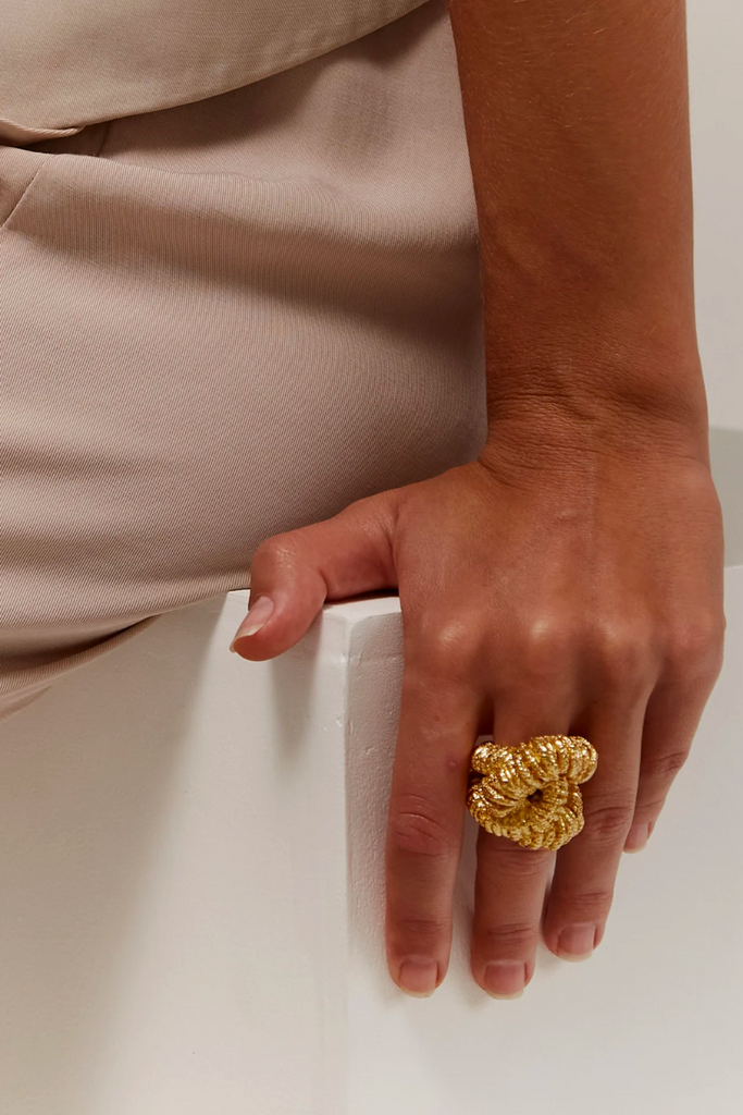 The Paola Sighinolfi Era Ring is a handcrafted 18kt gold plated statement piece with a unique grainy texture, intricate knot detailing, and an open band design, perfect to elevate any outfit.