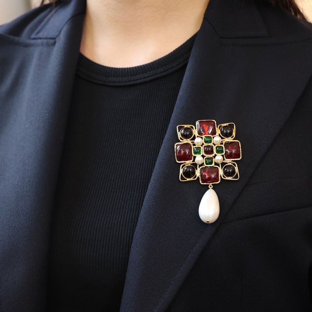 Person wearing a dark blazer with a decorative brooch featuring red, green, and gold colors.