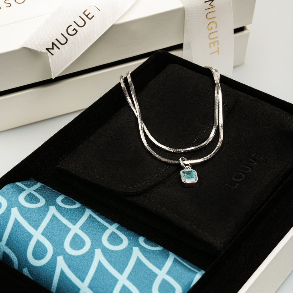 Silver necklace with a blue pendant on a black box with 'LOVE' branding, next to a white box with 'MAISON MUGUET' branding. Anniversary Limited Edition Gift Set by LOUVE 